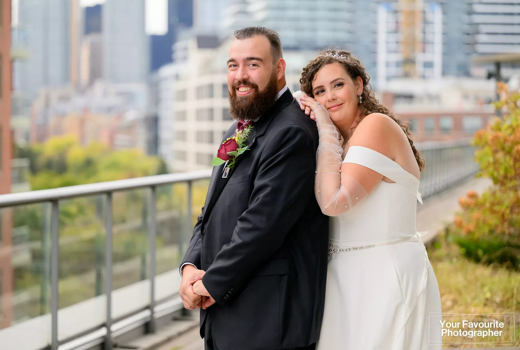 Bride leans against a groom as they pose on a rooftop patio with a garden and city in the background