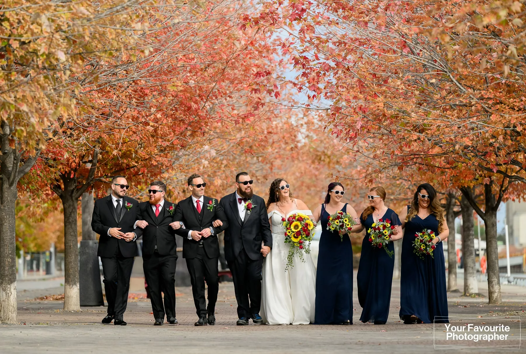 Wedding party wearing sunglasses walks down the Water's Edge Promenade in downtown Toronto, surrounded by fall foliage