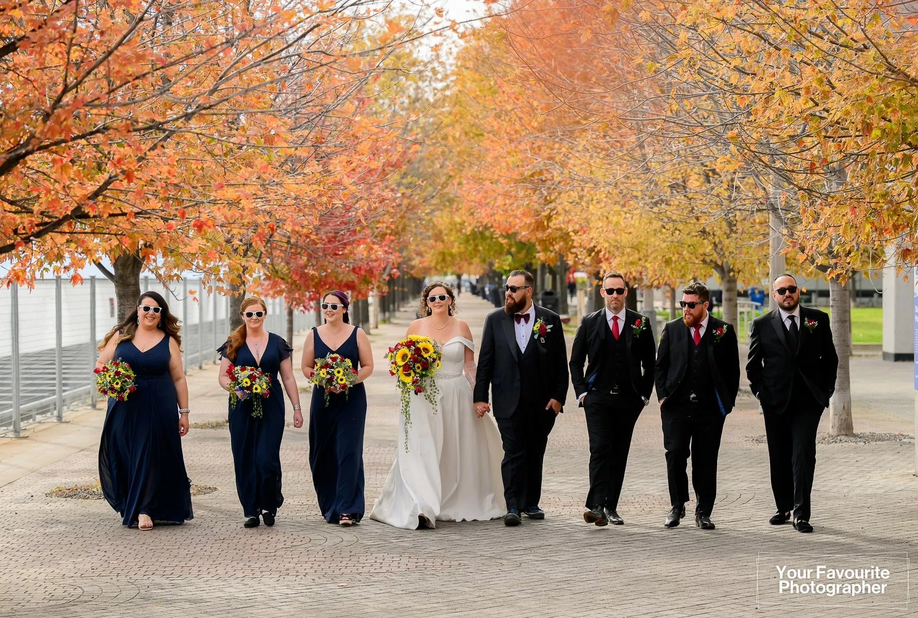 Wedding party walks down the Water's Edge Promenade in downtown Toronto, surrounded by fall foliage