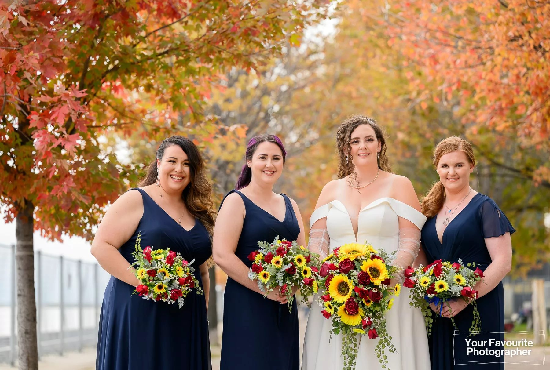 Bride and bridesmaids posing on the Water's Edge Promenade in front of Sherbourne Common in downtown Toronto. They are surrounded by colourful fall foliage on a bright day.