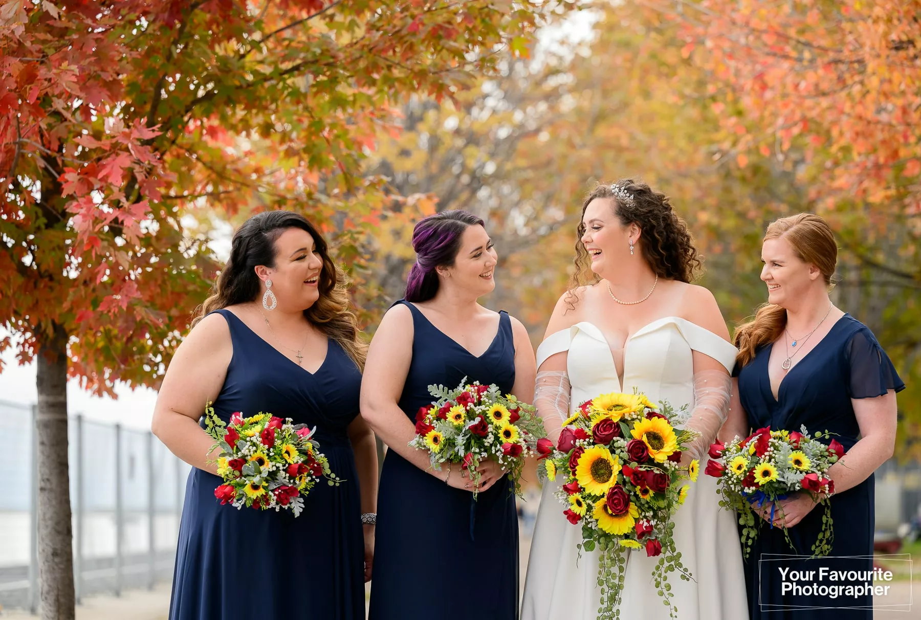 Bride and bridesmaids posing on the Water's Edge Promenade in front of Sherbourne Common in downtown Toronto. They are surrounded by colourful fall foliage on a bright day.