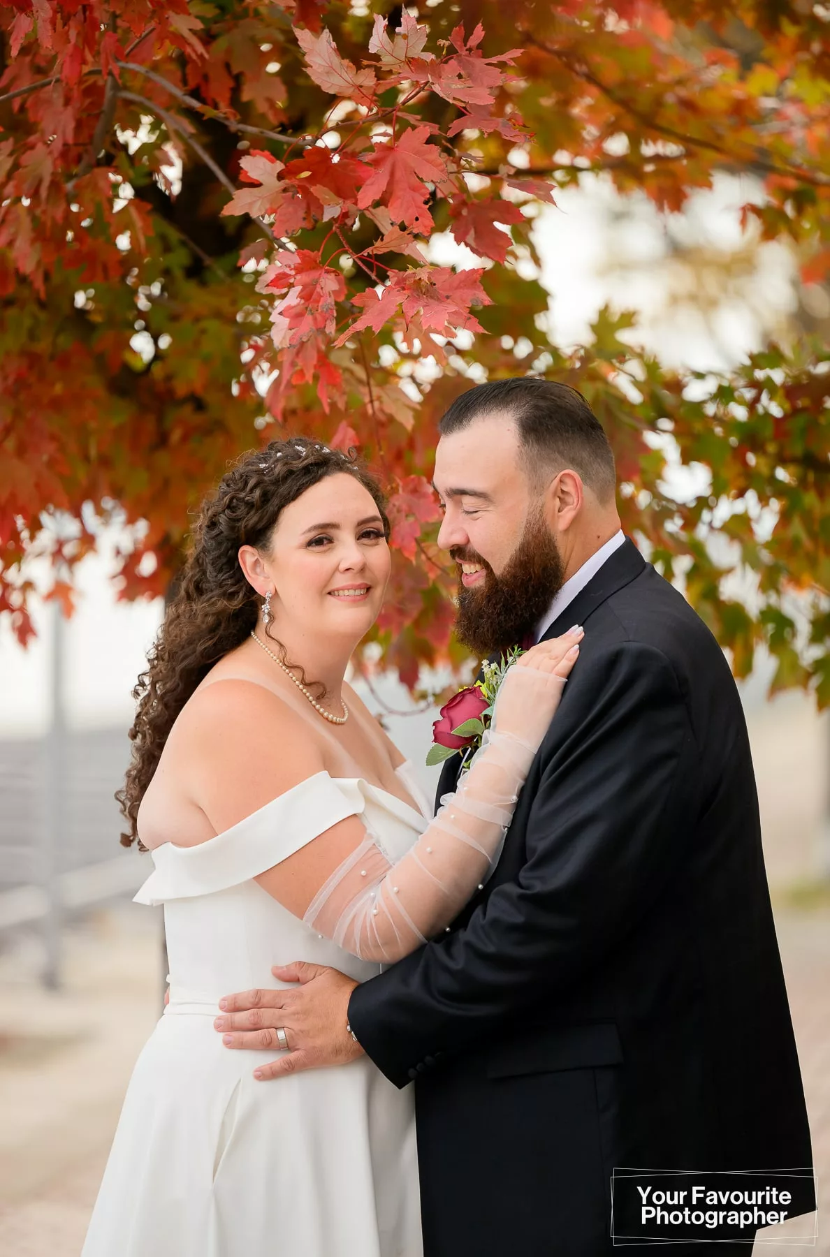 Bride and groom Emily and Niko pose, standing in front of colourful fall foliage on the Water's Edge Promenade in downtown Toronto