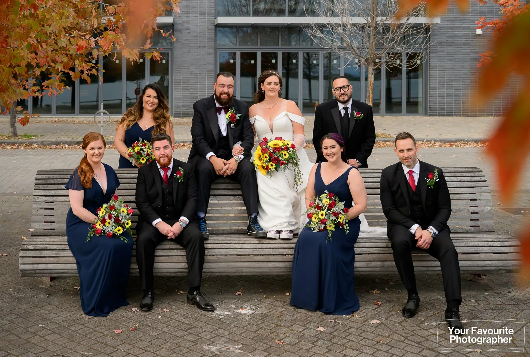 Bride and groom with their bridesmaids and groomsmen sit on a bench in downtown Toronto, near Sherbourne Common, on the Water's Edge Promenade