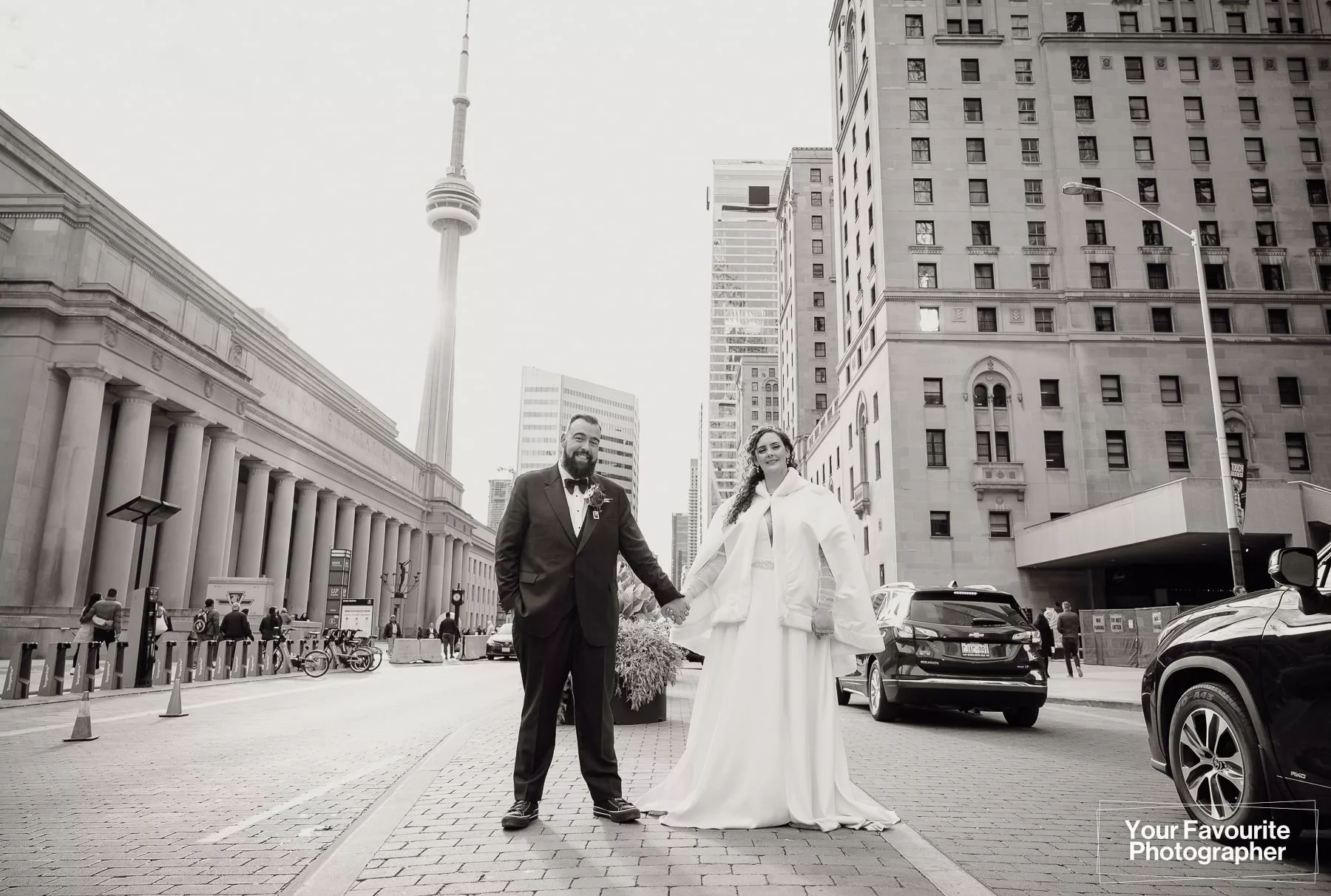 Black and white photo of a bride and groom posing in front of Union Station on Front St in downtown Toronto, with the Royal York hotel and CN Tower in the background