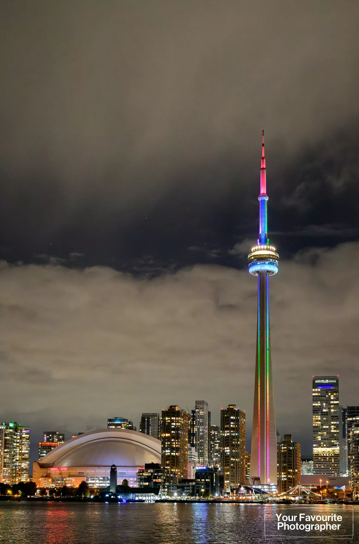 CN Tower lit up in rainbow colours pictured next to the SkyDome (Rogers Centre) on a cloudy night