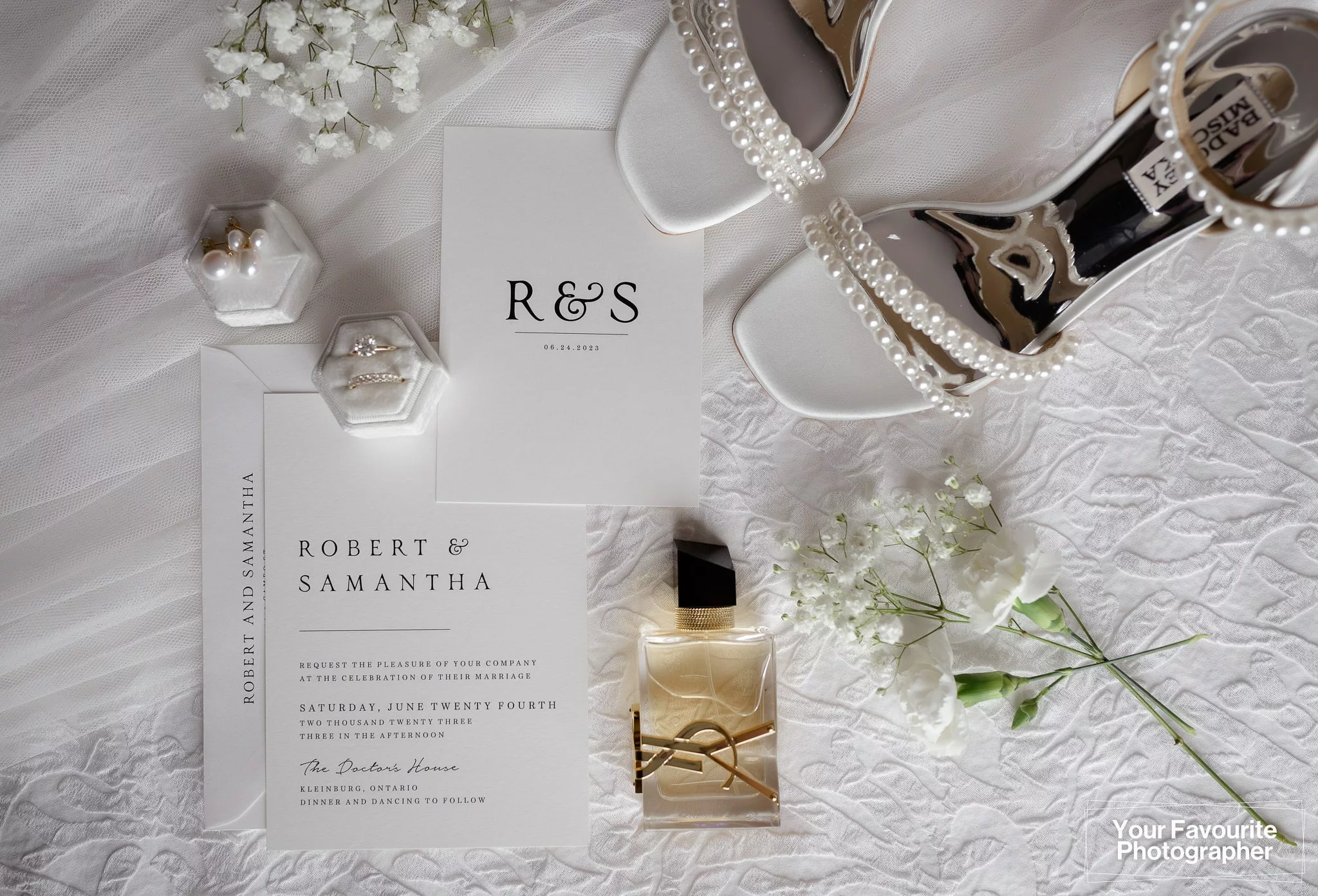 Flat lay of a bride's wedding accessories including YSL perfume, white flowers, a wedding invitation, wedding rings, and shoes