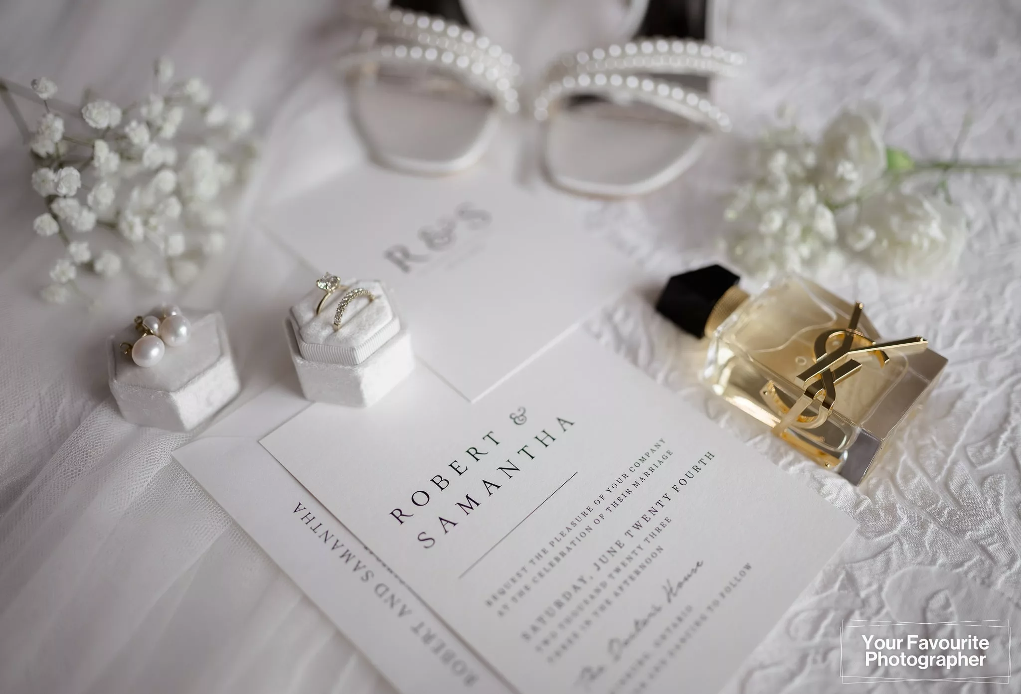 Flat lay of a bride's wedding accessories including YSL perfume, white flowers, a wedding invitation, wedding rings, and shoes