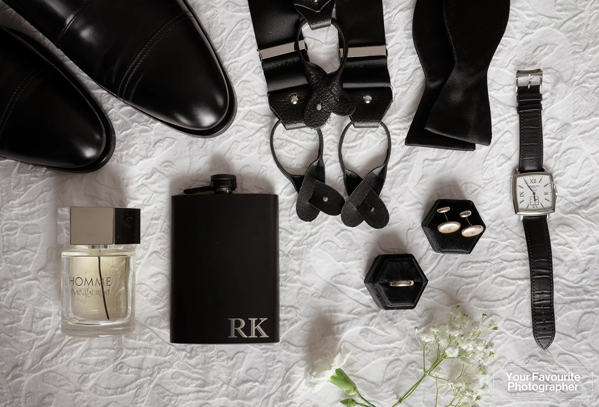 Flat lay of a groom's wedding accessories including YSL cologne, flask, wedding band, cufflinks, watch, suspenders, and shoes