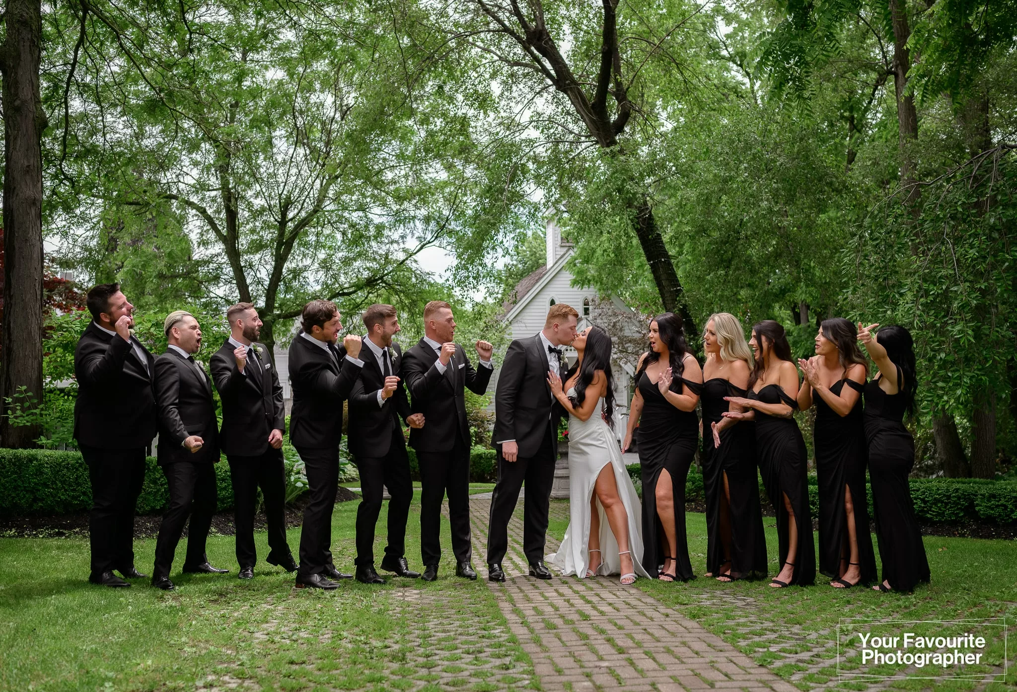 Formal photo of a wedding party of 13 people at The Doctor's House, all dressed in black and white outfits. The party celebrates as the couple shares a kiss.