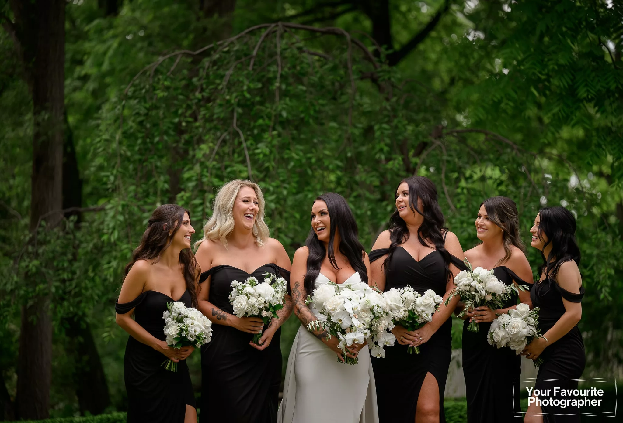 Bride with her bridesmaids in a black dresses and white flowers at The Doctor's House, standing in front of lush green trees