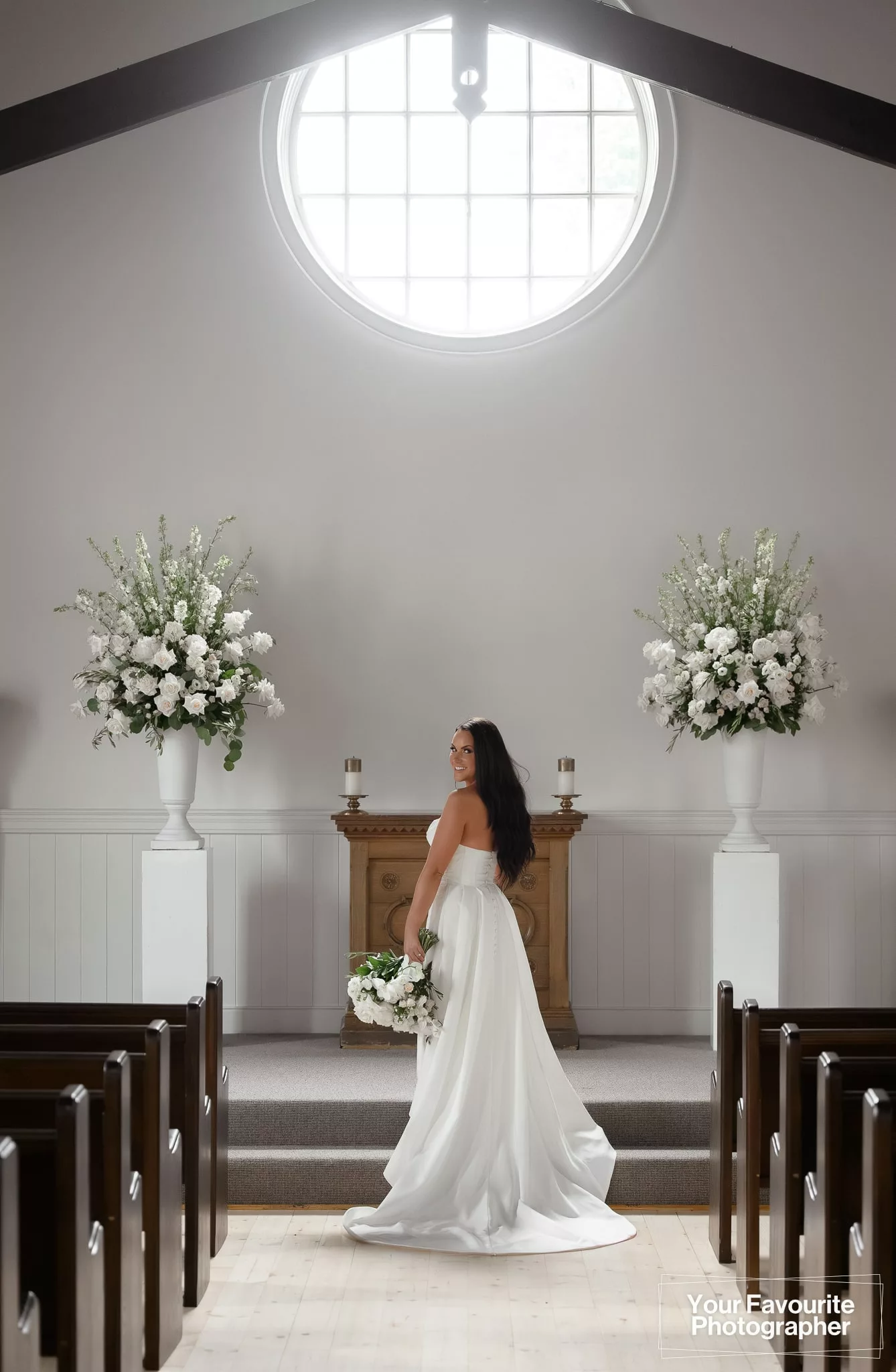 Bride standing alone at the altar with her bouquet, flanked by pedestals with white flowers